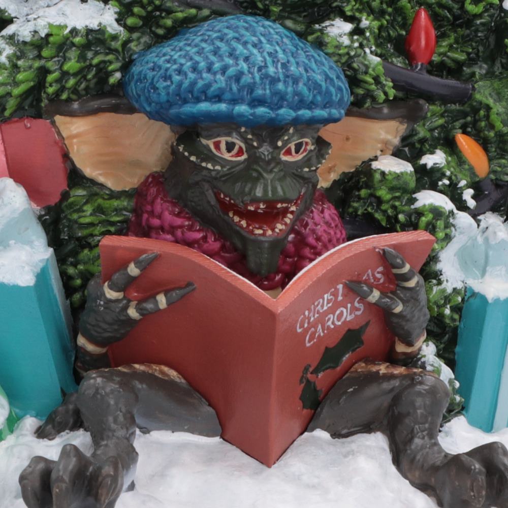 Mischievous Gremlins wearing a blue knit hat and pink scarf, sitting in snow and reading a red book that says 'Christmas Carols'.