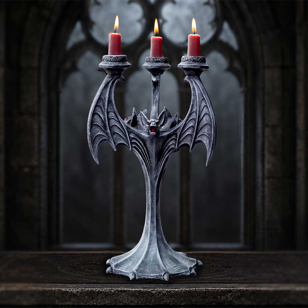 A black bat-shaped three-arm candelabra with detailed wing textures and a small red accent near the center,
holding three lit red candles, displayed on a dark wooden surface against a dark gothic arch background.